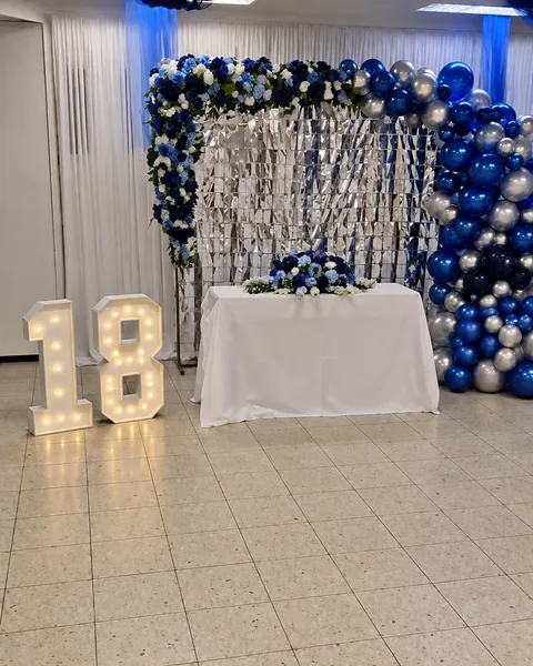 Koristeltu juhlaseinä: kukkakaari, hopeinen seinä, sini-hopeiset ilmapallot ja valaistut numerot 18.